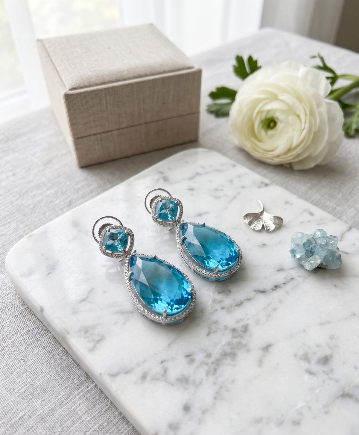 Blue teardrop earrings on a marble surface with a white flower and jewelry box in the background.