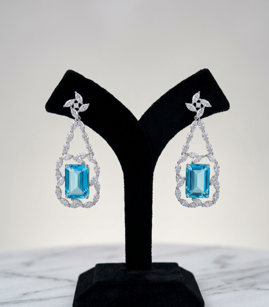 A pair of drop earrings displayed on a black jewelry stand against a light gray background. Each earring features a rectangular light blue gemstone surrounded by a halo of clear stones, suspended from a silver-toned floral connector.
