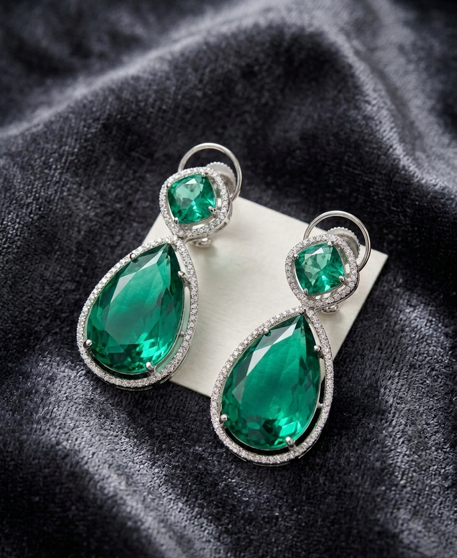 Pair of emerald green teardrop earrings with silver accents on a dark fabric background