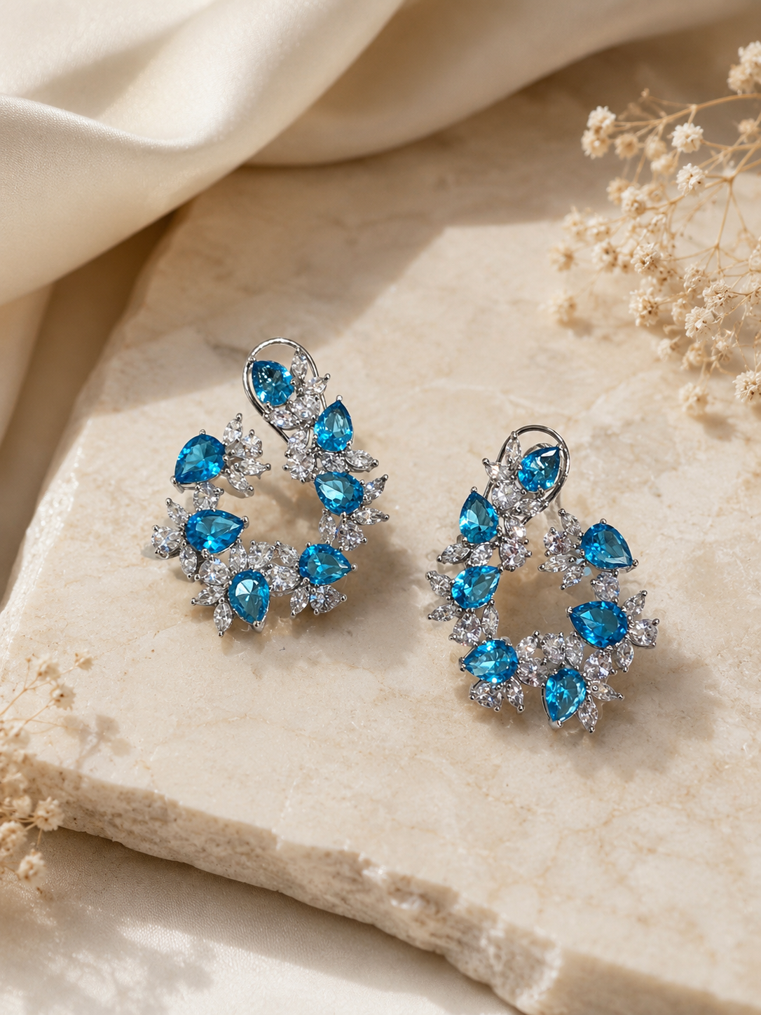 Pair of blue gemstone earrings on a beige stone surface with dried flowers.