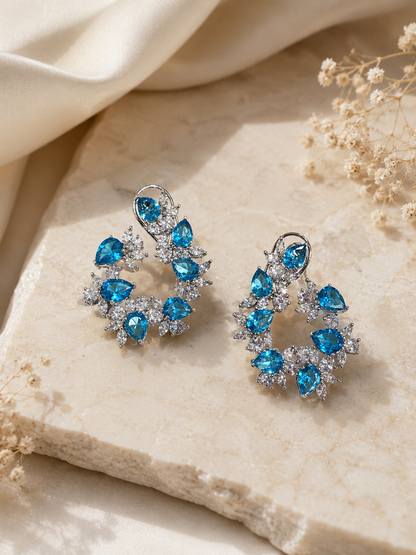 Pair of blue gemstone earrings on a beige stone surface with dried flowers.