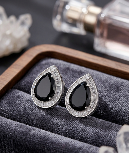 Teardrop-shaped black gemstone earrings with silver setting on a gray cushion, with perfume bottles in the background.