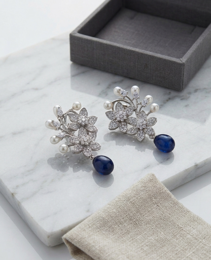 Pair of earrings with pearl and blue stone design on a marble surface with a gray box and beige cloth.