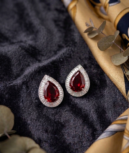 Teardrop-shaped red gemstone earrings on a dark fabric background with gold fabric and leaves.