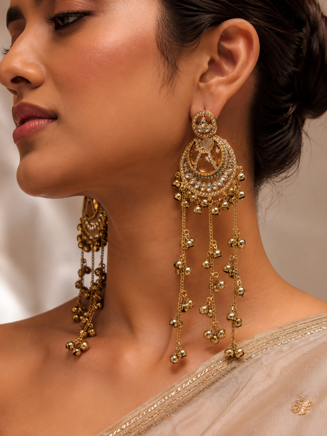 A close-up of a woman wearing ornate gold-toned traditional earrings with dangling chains and bead details, shown against a soft neutral background.