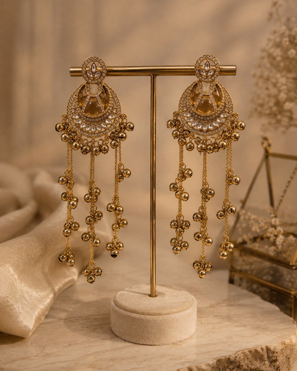 A pair of ornate gold-toned dangle earrings displayed on a gold stand, featuring intricate filigree, clear stones, and dangling beaded chains, set against a warm beige background with decorative fabric and dried floral elements.