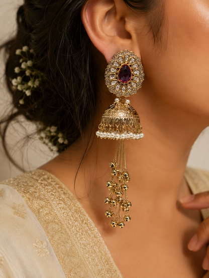 A close-up of a woman wearing an ornate gold-toned traditional Indian jhumka earring with a purple teardrop stone and white pearl accents.