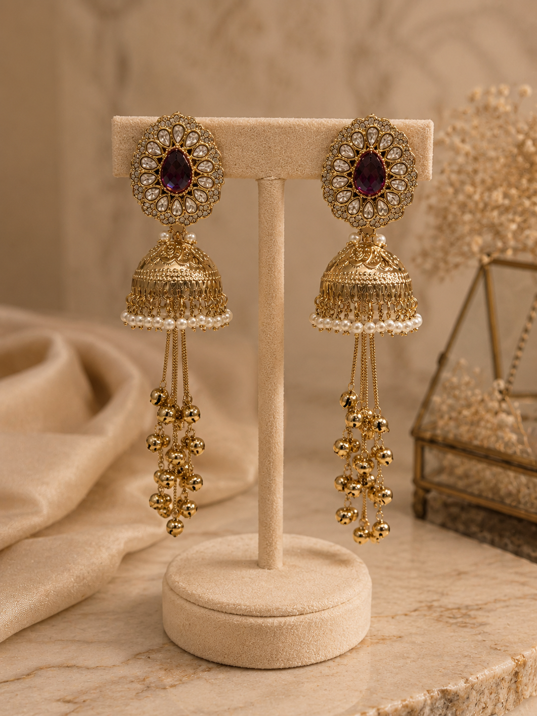 A pair of ornate gold-toned jhumka earrings with oval purple stones and dangling beaded chains, displayed on a beige jewelry stand against a warm neutral background.