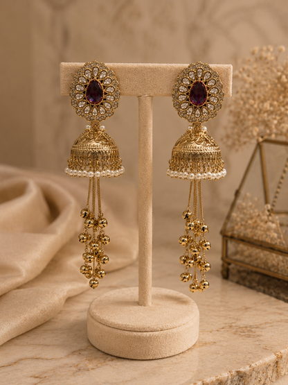 A pair of ornate gold-toned jhumka earrings with oval purple stones and dangling beaded chains, displayed on a beige jewelry stand against a warm neutral background.