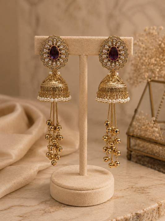 A pair of ornate gold-toned jhumka earrings with oval purple stones and dangling beaded chains, displayed on a beige jewelry stand against a warm neutral background.