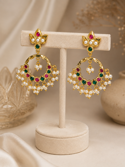 A pair of gold-toned dangle earrings with green and red stones and white pearl-like beads, displayed on a cream jewelry stand against a soft beige background.