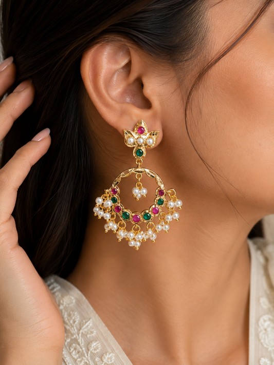 A close-up of a woman wearing a large gold-toned traditional Indian jhumka earring adorned with white pearls and red and green stones.