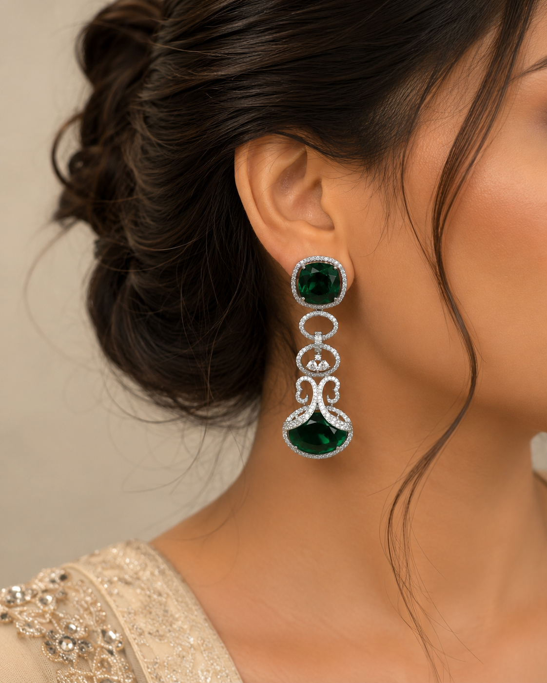 A close-up of a woman wearing a long, dangling statement earring. The earring features a round green gemstone at the top, a silver-toned decorative link chain with small clear stones, and a larger teardrop-shaped green gemstone at the bottom.