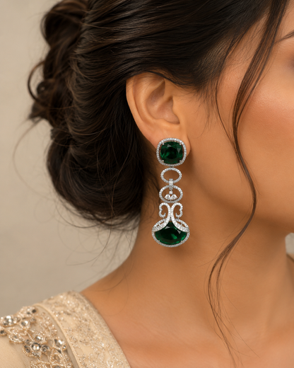 A close-up of a woman wearing a long, dangling statement earring. The earring features a round green gemstone at the top, a silver-toned decorative link chain with small clear stones, and a larger teardrop-shaped green gemstone at the bottom.