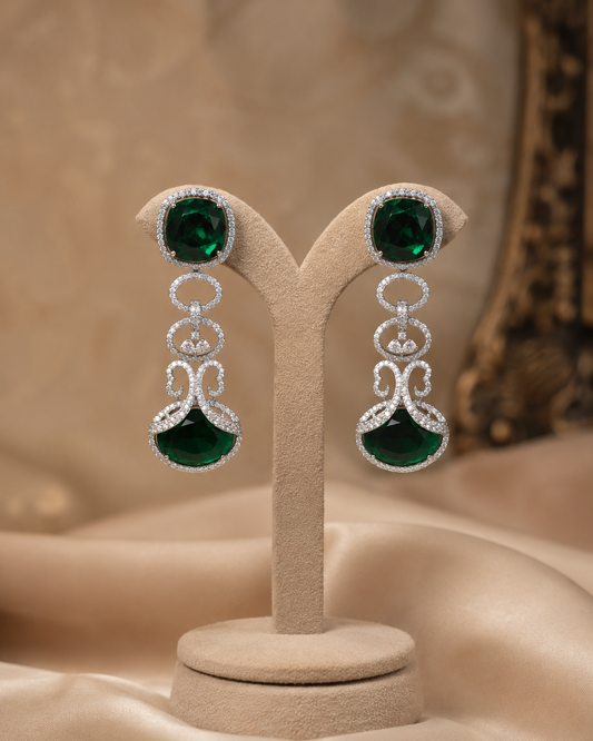 A pair of silver drop earrings with large oval green gemstones and smaller clear stones, displayed on a beige jewelry stand against a warm beige satin background.