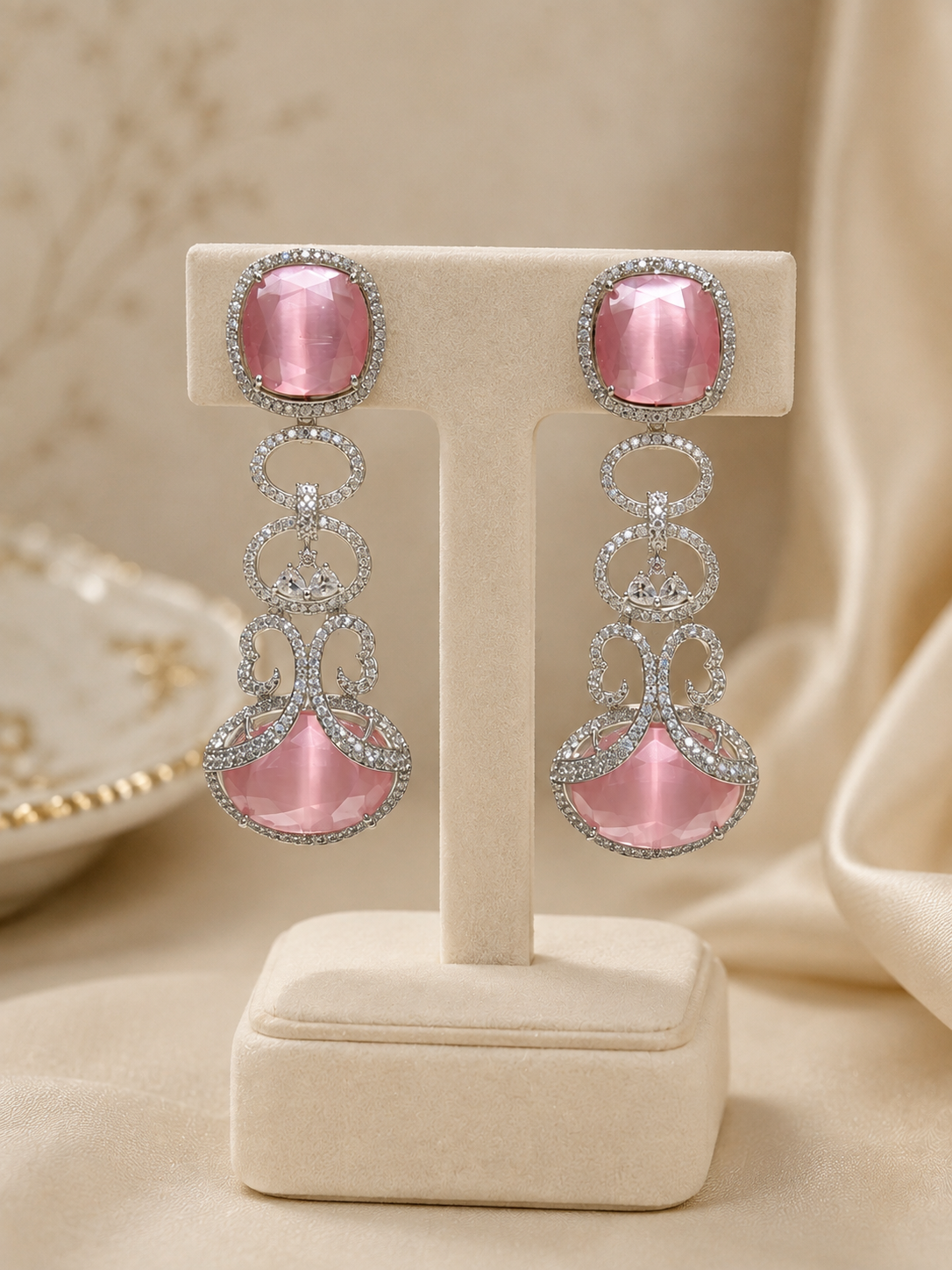 A pair of pink gemstone drop earrings with silver-toned metal settings and clear stones, displayed on a cream jewelry stand against a soft beige fabric background.
