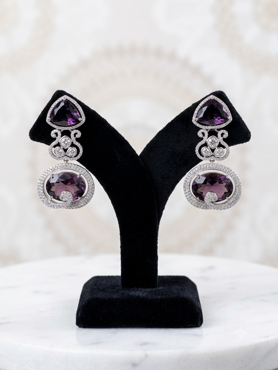 A pair of silver-toned drop earrings with triangular purple amethyst top stones and oval purple amethyst pendant stones, displayed on a black velvet jewelry stand over a white marble surface.