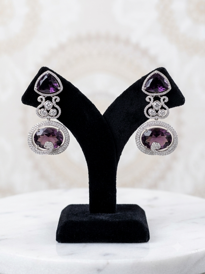 A pair of silver-toned drop earrings with triangular purple amethyst top stones and oval purple amethyst pendant stones, displayed on a black velvet jewelry stand over a white marble surface.