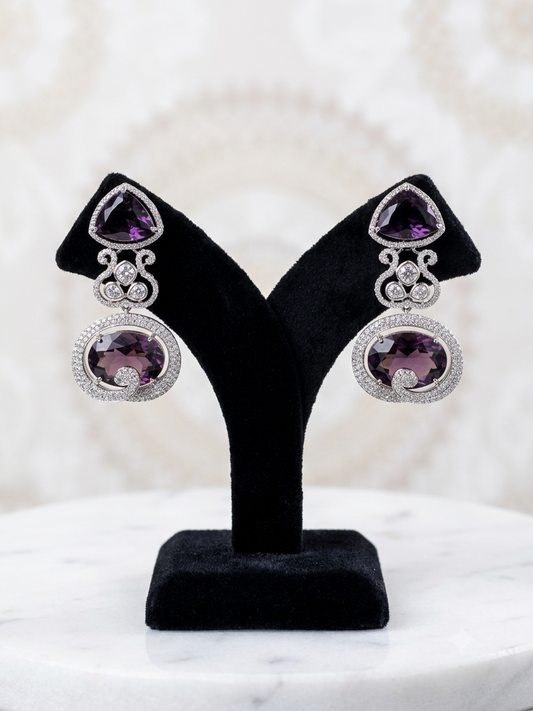 A pair of silver-toned drop earrings with triangular purple amethyst top stones and oval purple amethyst pendant stones, displayed on a black velvet jewelry stand over a white marble surface.