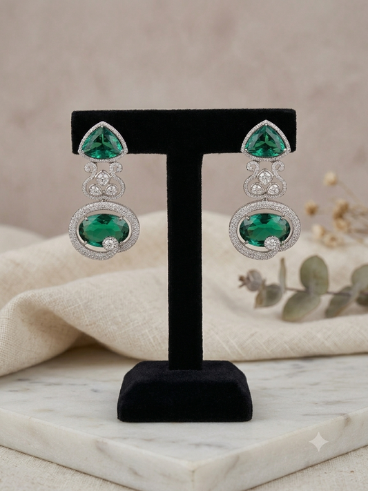 A pair of silver drop earrings with large green heart-shaped gemstones and smaller clear stones, displayed on a black T-stand over a cream fabric and marble surface.