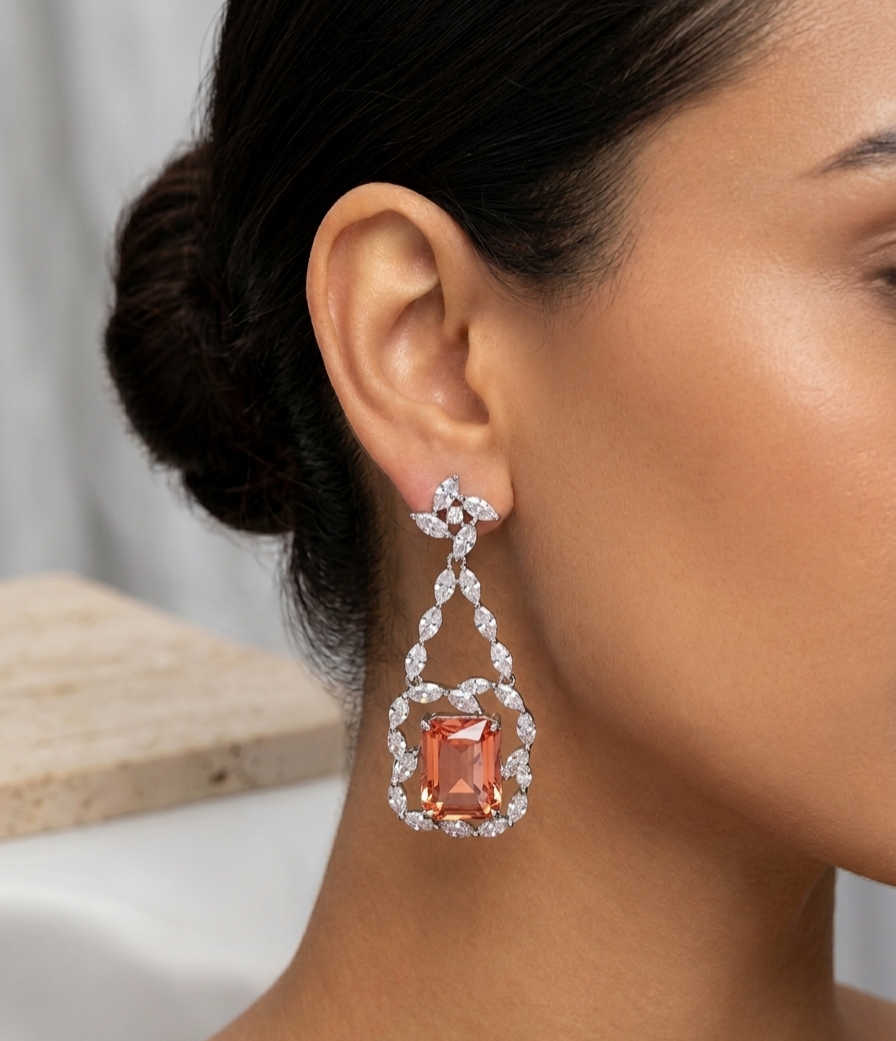A close-up of a woman's ear and neck wearing a dangling earring. The earring features a large rectangular peach-orange center stone surrounded by a halo of small clear stones, with additional clear stones forming a bow at the top and a vertical drop leading to the center stone.
