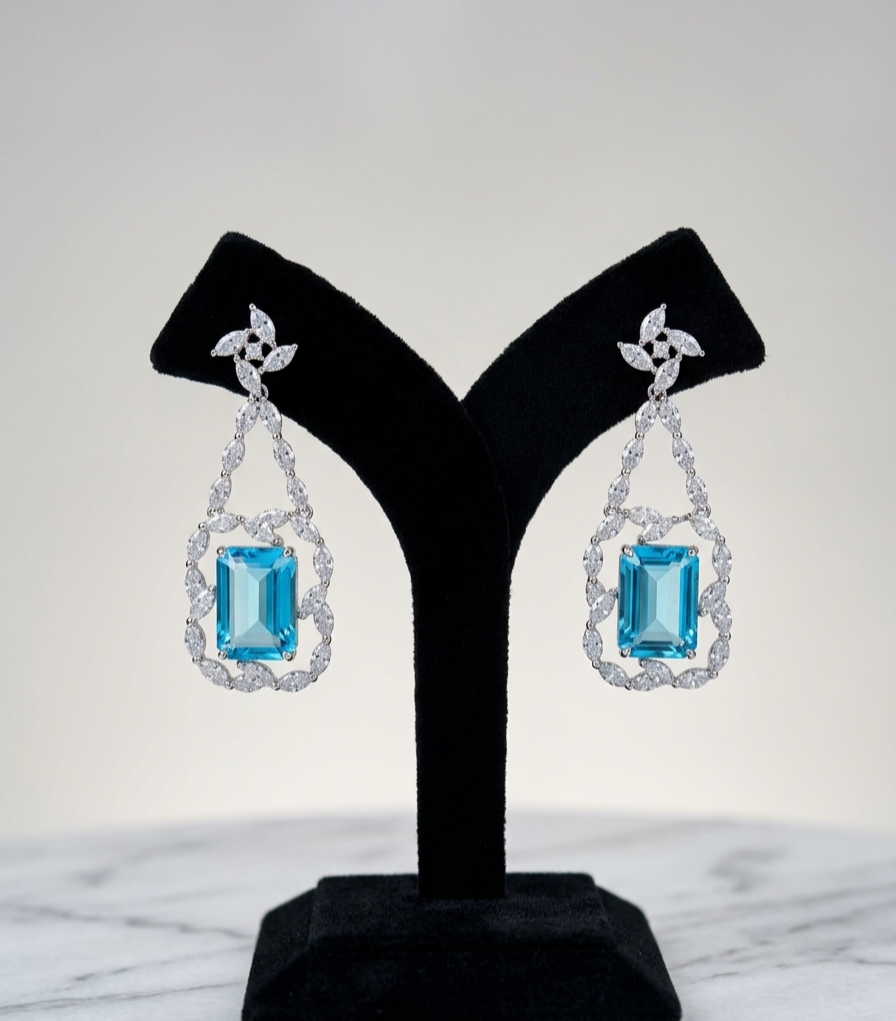 A pair of drop earrings displayed on a black jewelry stand against a light gray background. Each earring features a rectangular light blue gemstone surrounded by a halo of clear stones, suspended from a silver-toned floral connector.