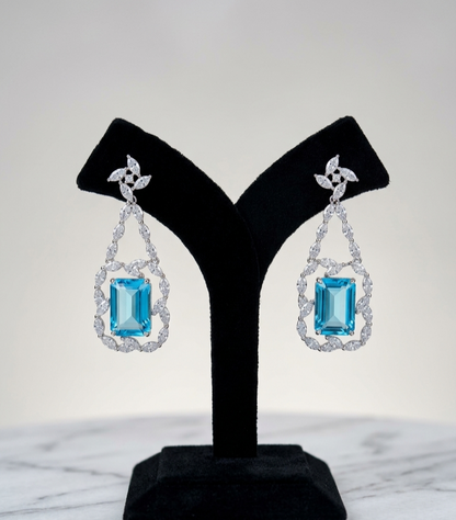 A pair of drop earrings displayed on a black jewelry stand against a light gray background. Each earring features a rectangular light blue gemstone surrounded by a halo of clear stones, suspended from a silver-toned floral connector.
