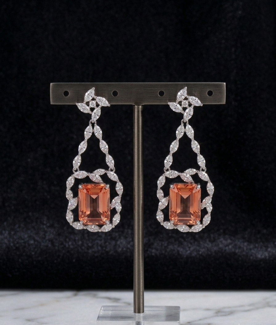 A pair of drop earrings displayed on a metal stand against a dark velvet background. Each earring features a rectangular peach-orange center stone surrounded by a silver-toned ornate frame set with small clear stones.