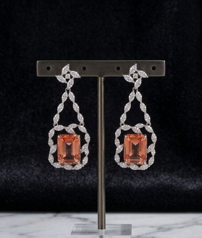 A pair of drop earrings displayed on a metal stand against a dark velvet background. Each earring features a rectangular peach-orange center stone surrounded by a silver-toned ornate frame set with small clear stones.