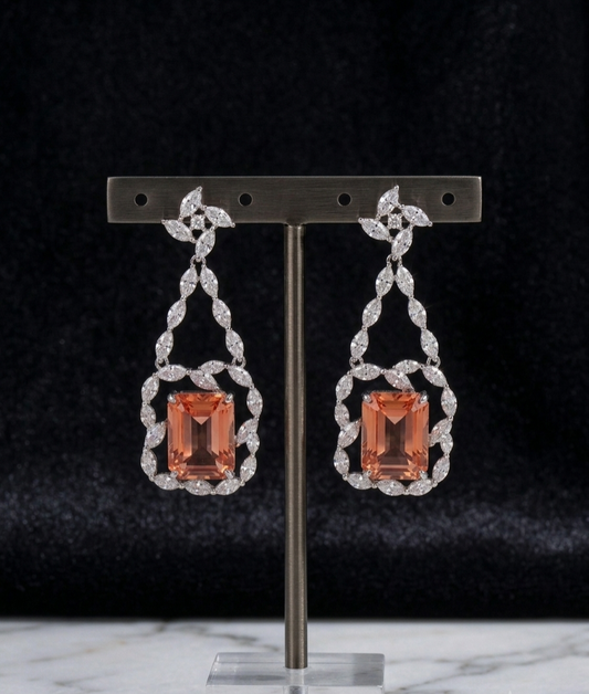 A pair of drop earrings displayed on a metal stand against a dark velvet background. Each earring features a rectangular peach-orange center stone surrounded by a silver-toned ornate frame set with small clear stones.
