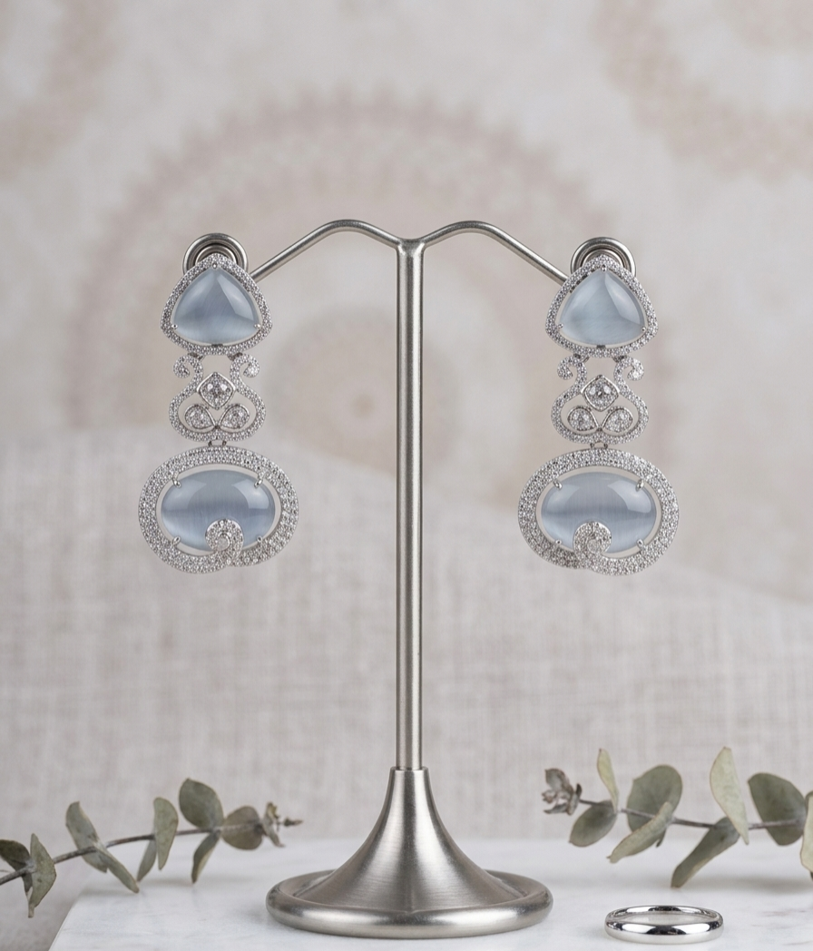 A pair of silver-toned dangle earrings with large pale blue oval stones and smaller clear stone accents, displayed on a silver stand against a soft beige textured backdrop with eucalyptus sprigs and a ring nearby.