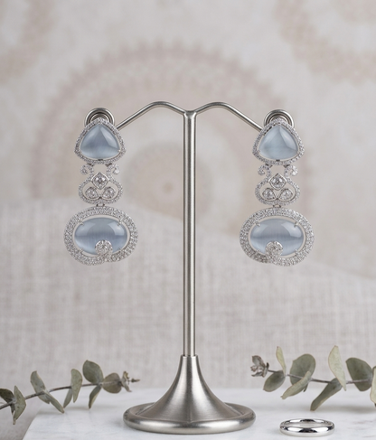 A pair of silver-toned dangle earrings with large pale blue oval stones and smaller clear stone accents, displayed on a silver stand against a soft beige textured backdrop with eucalyptus sprigs and a ring nearby.