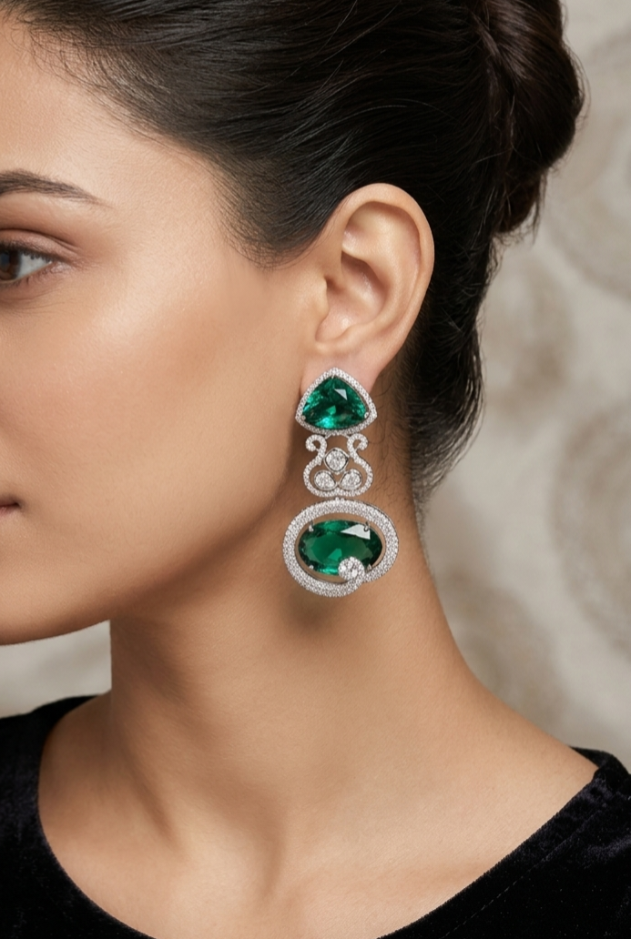 A close-up of a woman wearing a silver drop earring with two green gemstones and small clear accent stones.
