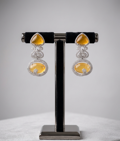 A pair of ornate dangle earrings displayed on a black velvet jewelry stand against a soft gray studio background. Each earring features a triangular amber-yellow top stone, a silver-toned filigree connector, and a larger oval amber-yellow drop stone surrounded by small clear stones.