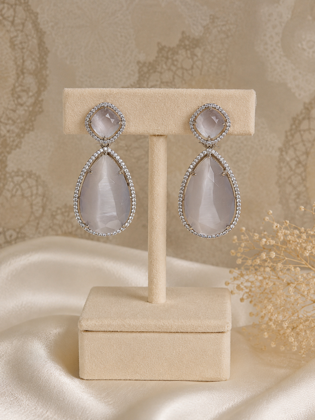 A pair of teardrop drop earrings with pale pinkish-white teardrop gemstones and oval round stones, set in silver-tone metal with small clear stones, displayed on a beige jewelry stand.
