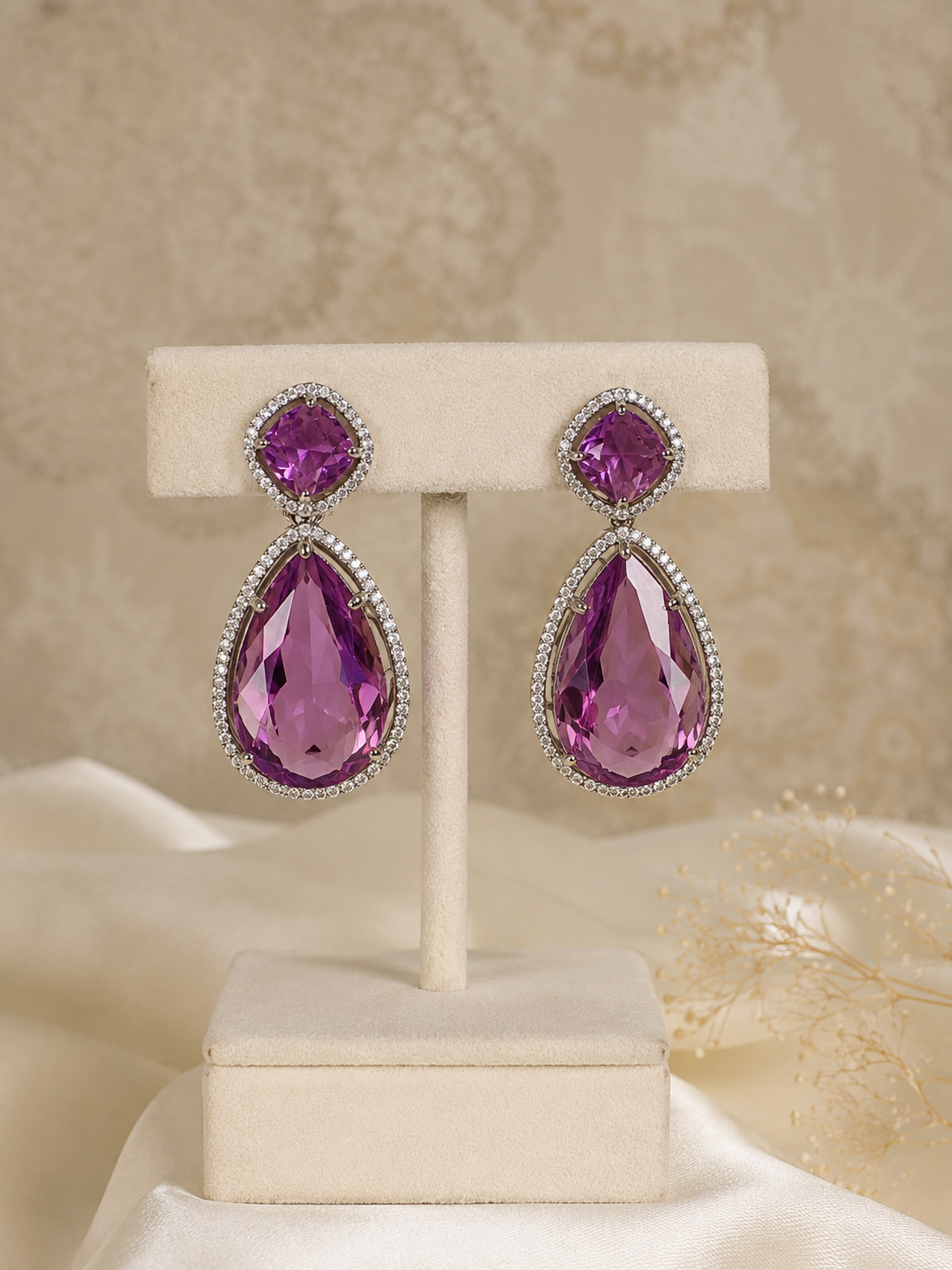 A pair of teardrop-shaped drop earrings with large faceted purple gemstones in silver settings, displayed on a cream jewelry stand against a soft beige patterned background.