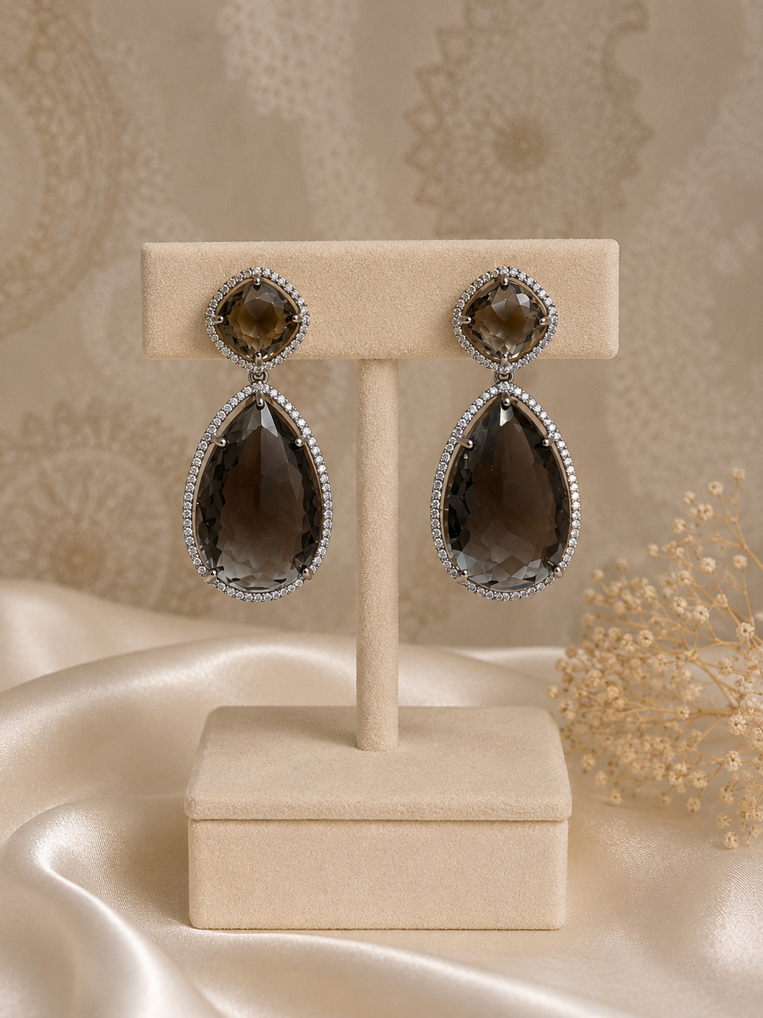 A pair of teardrop-shaped drop earrings with large smoky brown gemstones and small clear stones around the top round stones, displayed on a beige jewelry stand against a warm beige patterned backdrop with satin fabric and dried flowers.